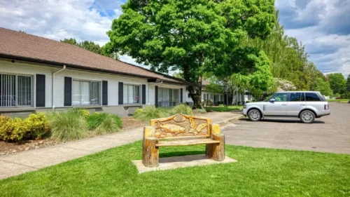 A white brick building with a brown roof and a walkway, with a carved wooden bench and a large tree in the foreground